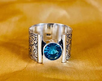 Puede incluir: Un anillo de plata con una gran gema azul brillante y redonda en el centro. La banda tiene un diseño grabado y ornamentado en los laterales. Dos barras verticales de plata flanquean la gema, añadiendo un toque moderno.