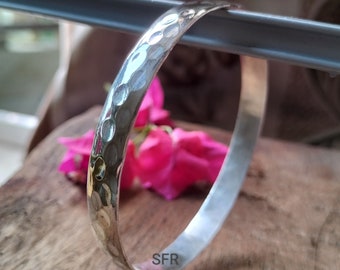 May include: A silver hammered bangle bracelet with a smooth, polished interior. The bracelet is marked with the letters "SFR".