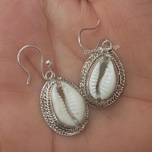 May include: Silver earrings featuring a white cowrie shell design. The earrings have a delicate, intricate design and are perfect for adding a touch of elegance to any outfit.