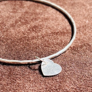 Puede incluir: Un brazalete plateado con un colgante en forma de corazón. El brazalete es circular y tiene una superficie lisa y reflectante. El corazón es plano y cuelga de un pequeño lazo. El fondo es una superficie marrón texturizada.