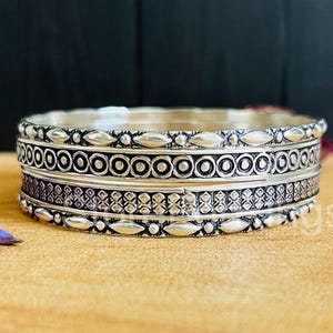 May include: A silver bangle bracelet with intricate geometric designs. The bracelet is made of multiple bands, each with a different pattern.