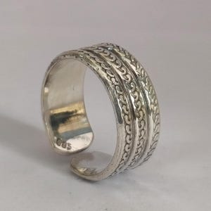 May include: A silver adjustable ring with an ornate, textured design. The ring features three bands of repeating patterns. The open design allows for size adjustment. The ring is on a white background.