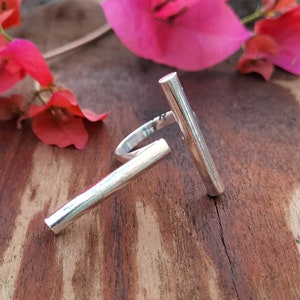 Puede incluir: Anillo de plata con dos barras verticales, una encima de la otra, formando una T. El anillo está sobre una superficie de madera.