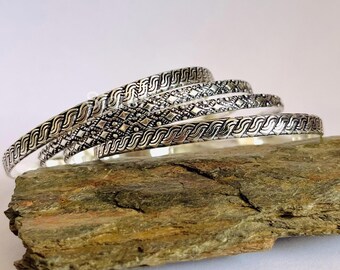 May include: Four silver bangles with intricate geometric designs. The bangles are stacked on top of each other and are resting on a rough, brown rock.