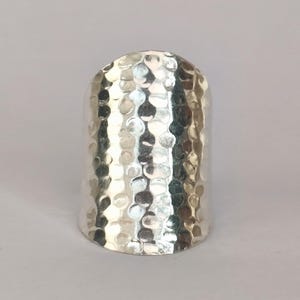 Peut inclure: Une large bague argentée à texture martelée. La bague a un dessus arrondi et un intérieur lisse. La texture martelée crée un motif de petites indentations irrégulières sur la surface. Un bijou audacieux.