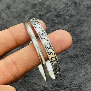 May include: Two silver bangles. One is plain, the other is decorated with a repeating pattern of stars and crescent moons. The bangles are held by a hand against a dark background. The jewelry is likely made of sterling silver.