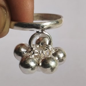 Puede incluir: Un anillo plateado con un grupo de pequeños adornos redondos colgantes. El anillo está sujeto por un dedo. Los adornos colgantes son esféricos y brillantes, reflejando la luz. El anillo parece estar hecho de metal brillante.
