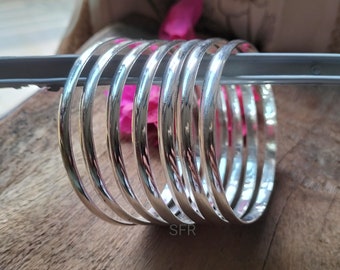 May include: A collection of shiny, silver bangles. The bracelets are round and smooth, reflecting light. The bangles are stacked together, resting on a wooden surface. The image has a soft focus background with pink flowers.