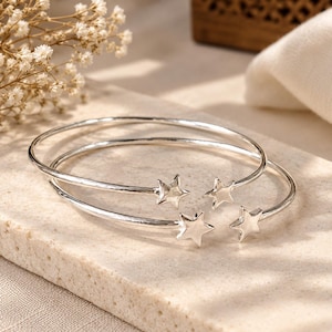 May include: Two silver-toned open cuff bracelets, each featuring a polished star at both ends. The bracelets have a smooth, rounded band design and are crafted from a reflective metallic material, showcasing a simple and clean aesthetic.