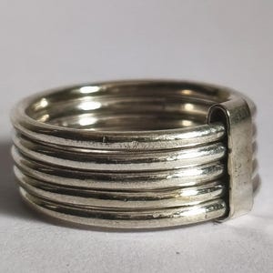 Puede incluir: Anillo de plata con un diseño único. El anillo presenta múltiples bandas apiladas, creando una apariencia texturizada. Las bandas están conectadas por una barra vertical en un lado. El anillo está hecho de un material metálico brillante.