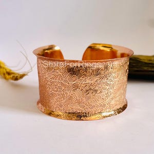 Könnte beinhalten: Ein Kupfer-Armreif mit einem komplizierten Blumenmuster. Der Armreif ist breit und hat eine abgerundete Form.