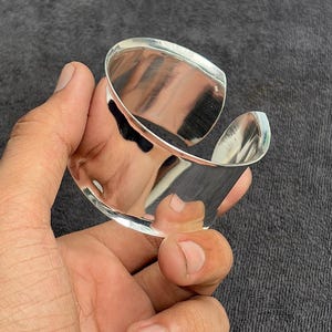 Könnte beinhalten: Ein poliertes silbernes Armreif-Armband mit geschwungenem, offenem Design. Das Armband reflektiert das Licht und erzeugt ein helles, glänzendes Aussehen. Der Schmuck wird in einer Hand vor einem dunkelgrauen Hintergrund gehalten.