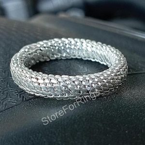 Può includere: Un anello d'argento con un design intrecciato. L'anello è realizzato con una serie di piccoli pezzi intrecciati che creano una superficie testurizzata.