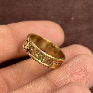 May include: A gold ring with a textured band featuring a repeating pattern of small, raised circles. The ring has a polished finish and a slightly curved design. The ring is shown on a person's finger.