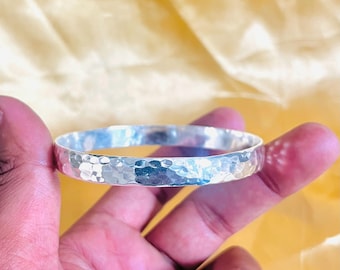 May include: A hammered silver bangle bracelet. The bracelet has a textured surface with a series of small, irregular indentations. The bangle is held in a hand against a yellow background.