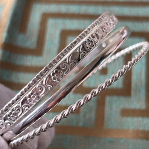 Op de afbeelding: Een set van drie zilveren armbanden. Eén armband heeft een bloemmotief, een andere is glad en de derde heeft een gedraaid touwontwerp. De armbanden worden gepresenteerd tegen een achtergrond met patronen.