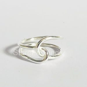 Op de afbeelding: Een zilverkleurige ring met een uniek, verweven ontwerp. De ring heeft twee banden die over elkaar lussen en kruisen, waardoor een open, knoopachtig uiterlijk ontstaat. De ring is gemaakt van een glanzend, reflecterend metaal.