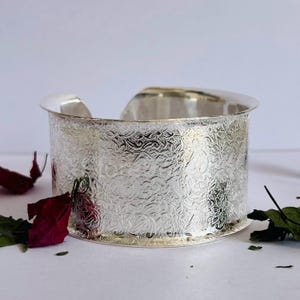 May include: A silver bangle bracelet with an intricate floral design. The bracelet is wide and has a smooth, polished finish.