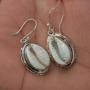 May include: Silver drop earrings with white cowrie shells. The shells are set in an oval frame with a decorative edge. The earrings have a simple hook closure, suitable for everyday wear or special occasions.