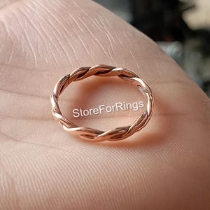 May include: A close-up of a rose gold braided ring. The ring is on a hand, but the hand is not visible in the image. The ring is simple and elegant.