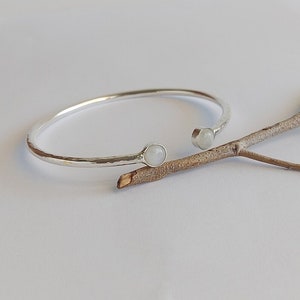 May include: A silver bangle bracelet with two white moonstone accents. The bracelet is hammered and has a slightly open design.