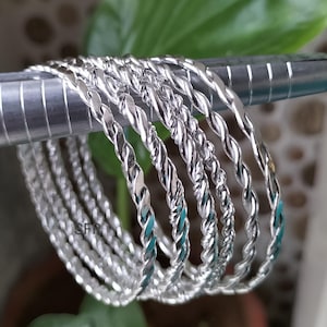 May include: A set of silver bangles with a twisted rope pattern. The bangles are displayed on a silver bar, with a green leafy backdrop. The shiny metal bangles reflect light, creating a bright appearance.