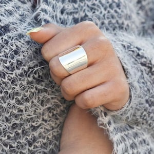 May include: A broad, polished silver ring with a rectangular face. The ring is worn on a finger, reflecting light. The background is a soft, textured grey sweater.