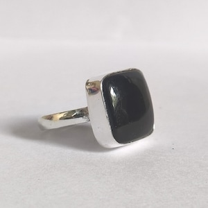 May include: A silver ring featuring a square, black gemstone. The stone is set in a silver bezel, and the band has a simple, polished design. The ring is displayed on a white surface.