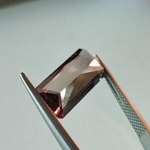 May include: A dark red gemstone with a rectangular cut, held by tweezers.