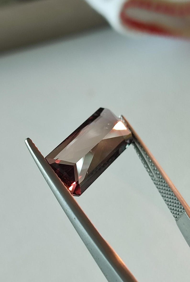 May include: A dark red gemstone with a rectangular cut, held by tweezers.