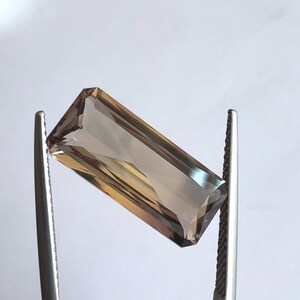 May include: A rectangular, faceted gemstone with a brown and purple hue. The gemstone is held in place by two metal tweezers.