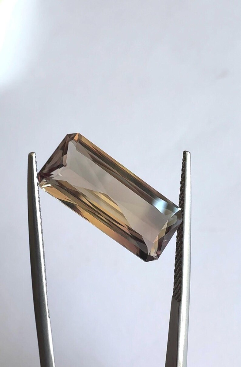 May include: A rectangular, faceted gemstone with a brown and purple hue. The gemstone is held in place by two metal tweezers.