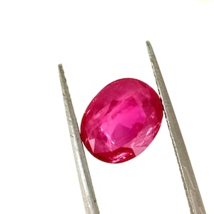 Könnte beinhalten: Ein leuchtend rosa ovaler Rubin-Edelstein, der von einer Pinzette gehalten wird.