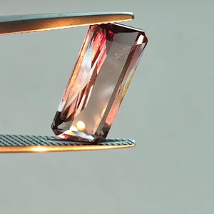May include: A pink and purple gemstone with a rectangular shape is held by a pair of tweezers.