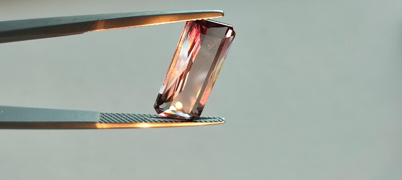 May include: A pink and purple gemstone with a rectangular shape is held by a pair of tweezers.