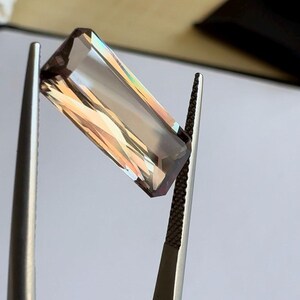 May include: A rectangular, faceted gemstone with a brown and iridescent color held by tweezers.