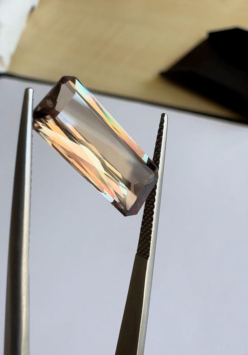 May include: A rectangular, faceted gemstone with a brown and iridescent color held by tweezers.