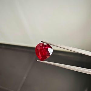 以下が含まれることがあります： ハート型の深紅色の宝石が銀色のピンセットで挟まれています。宝石のファセットが光を捉え、明るいハイライトを生み出しています。背景はグレーと白の柔らかなグラデーションです。