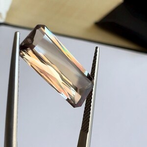 May include: A rectangular, faceted gemstone with a light brown color and iridescent rainbow sheen. The gemstone is held by tweezers.