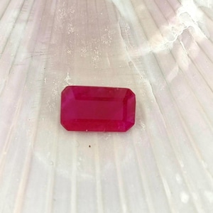 May include: A vibrant, deep red gemstone with a rectangular, emerald cut. The gemstone is set against a white, textured background.