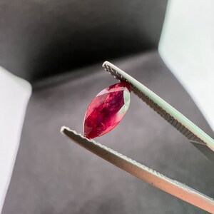 May include: A deep red marquise-shaped gemstone held by tweezers.