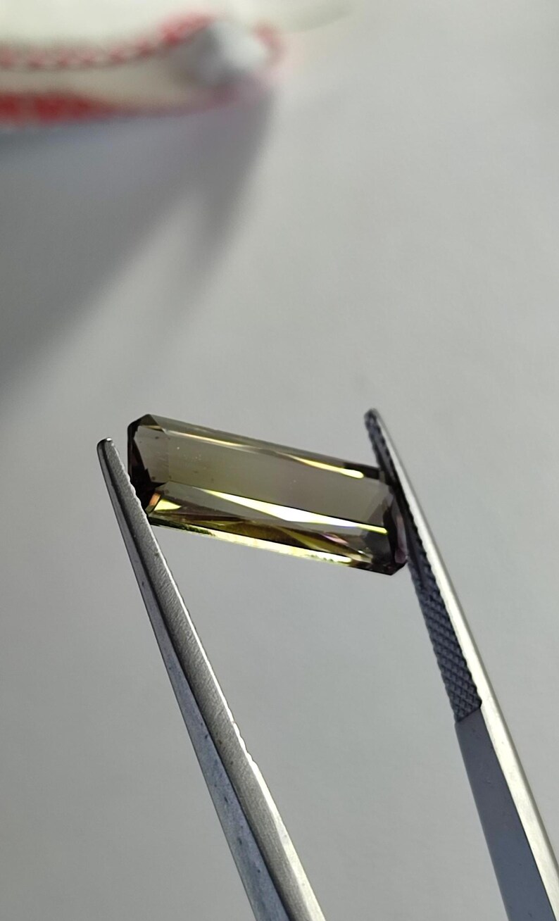 May include: A faceted, rectangular, brown and green gemstone held by tweezers.