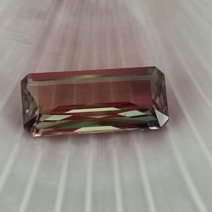 May include: A rectangular, faceted gemstone with a green, brown, and red color gradient. The gemstone is set against a white, textured background.