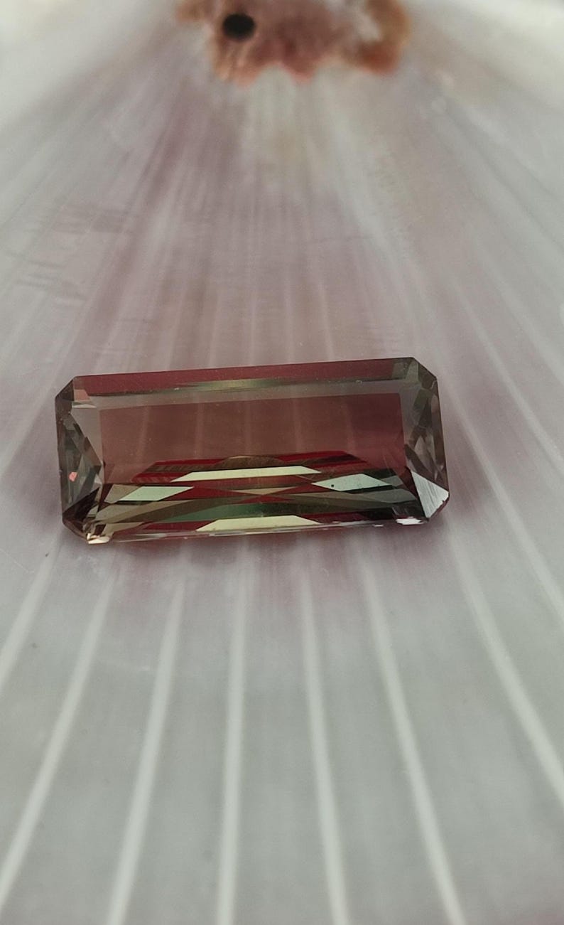 May include: A rectangular, faceted gemstone with a green, brown, and red color gradient. The gemstone is set against a white, textured background.