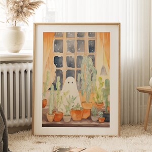 May include: Watercolour print of a friendly ghost surrounded by potted cacti and succulents in a window. The artwork features a warm colour palette with orange curtains and a night sky visible. The framed art piece is displayed on a fluffy rug.