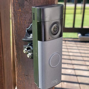 May include: A black and silver video doorbell with a camera lens at the top and a circular button in the centre. The word ring is printed in white at the bottom. It is mounted to a wooden post using a black metal bracket with visible screws.