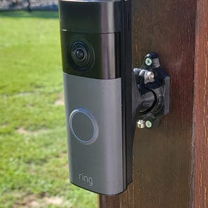 May include: A black and silver Ring video doorbell mounted to a dark brown wooden post using a black 3D-printed adjustable bracket. The device features a circular camera lens at the top, a central button, and the word ring printed at the bottom.