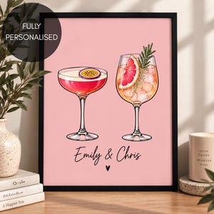 May include: A framed wall art print on a light pink background featuring two cocktail glasses. The left glass contains a pink drink with a passion fruit slice, and the right glass holds a drink with ice, a grapefruit slice, and a rosemary sprig.