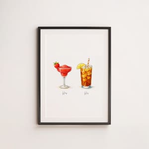 Op de afbeelding: Een ingelijste print met aquarel illustraties van twee cocktails. Eén is een rode aardbeienmargarita in een glas met steel, en de andere is een ijsthee met een citroenschijfje en een rietje. De woorden "Hers" en "His" staan eronder.