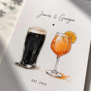 Puede incluir: Una ilustración en acuarela sobre papel texturizado que muestra un vaso de pinta con cerveza negra y una copa con un cóctel naranja y una rodaja de naranja. El texto dice James & Georgia con un pequeño corazón y la fecha EST. 2022 debajo.